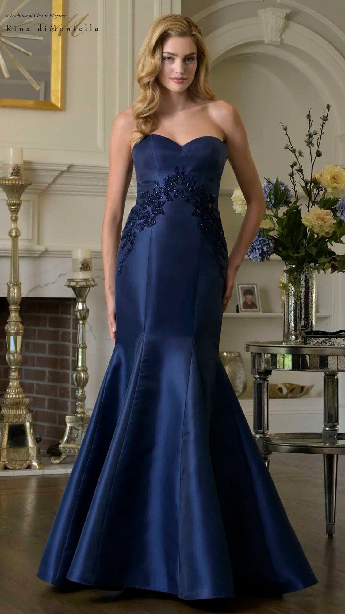 A woman poses indoors in front of a fireplace and floral arrangement, wearing the Long Formal Dress: Rina diMontella RD3420—a strapless, navy blue, floor-length gown with lace bodice detailing.